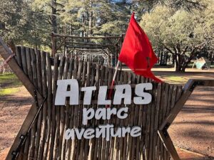 The rustic wooden entrance sign of Atlas Parc Aventure in the Ifrane cedar forest, featuring a Moroccan flag and the Forêt de Moudmane in the background.